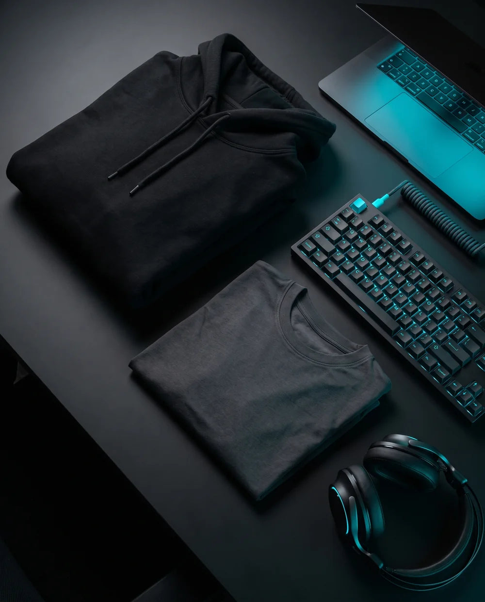 Black hoodie and tee flat-lay on a hacker desk with cyan-lit keyboard
